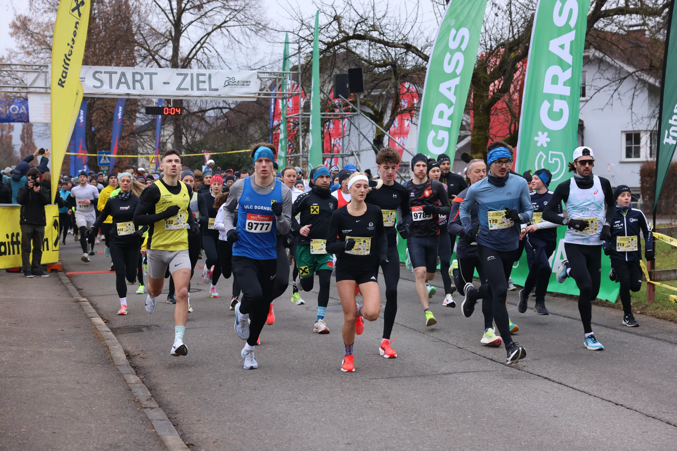 Eine Gruppe von Läufern beim Start eines Wettlaufs, mit einem Banner 'START-ZIEL' im Hintergrund. Teilnehmer tragen Sportkleidung und starten energisch auf einer Straße.